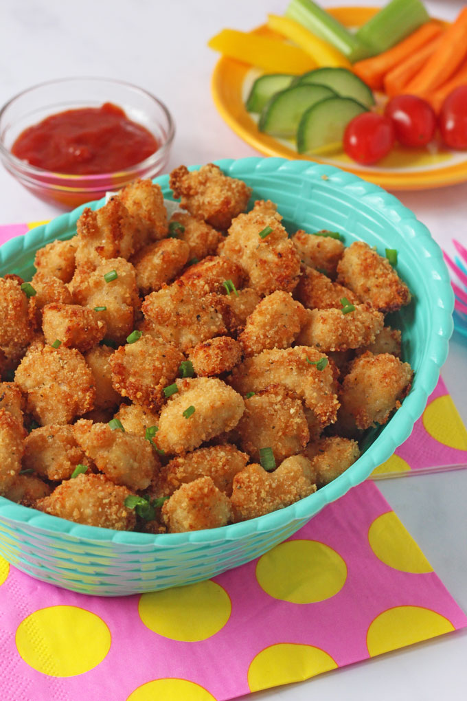 Crispy Baked Popcorn Chicken in a blue bowl with a selection of chopped vegetables in the background