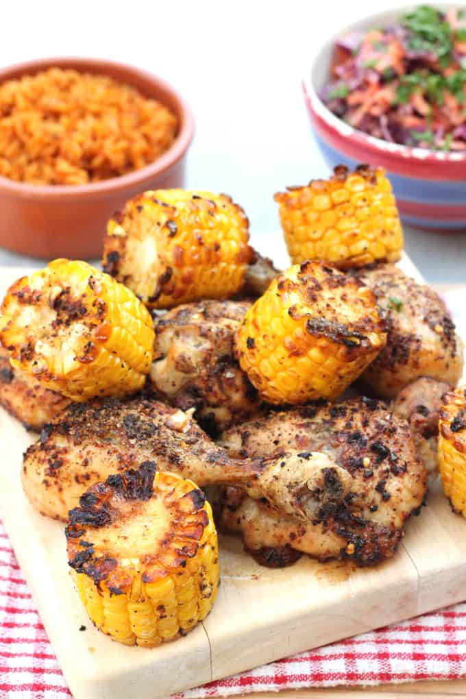 Spicy peri peri chicken thighs and chicken drumsticks and corn on the cob on a wooden chopping board with a bowl of spicy rice and a bowl of coleslaw in the background