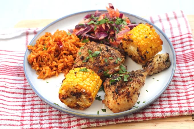 peri peri chicken thighs and chicken drumsticks served with spicy rice, coleslaw and corn on the cob on a white plate