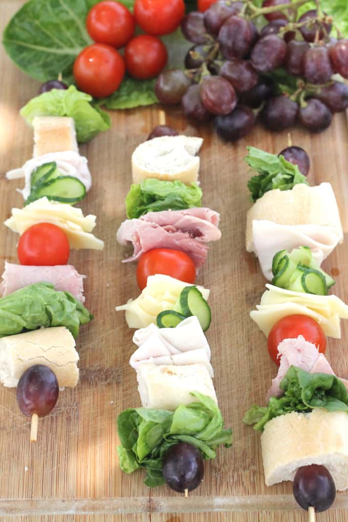 sandwich skewers on a wooden chopping board with black grapes, tomatoes and lettuce in the background.