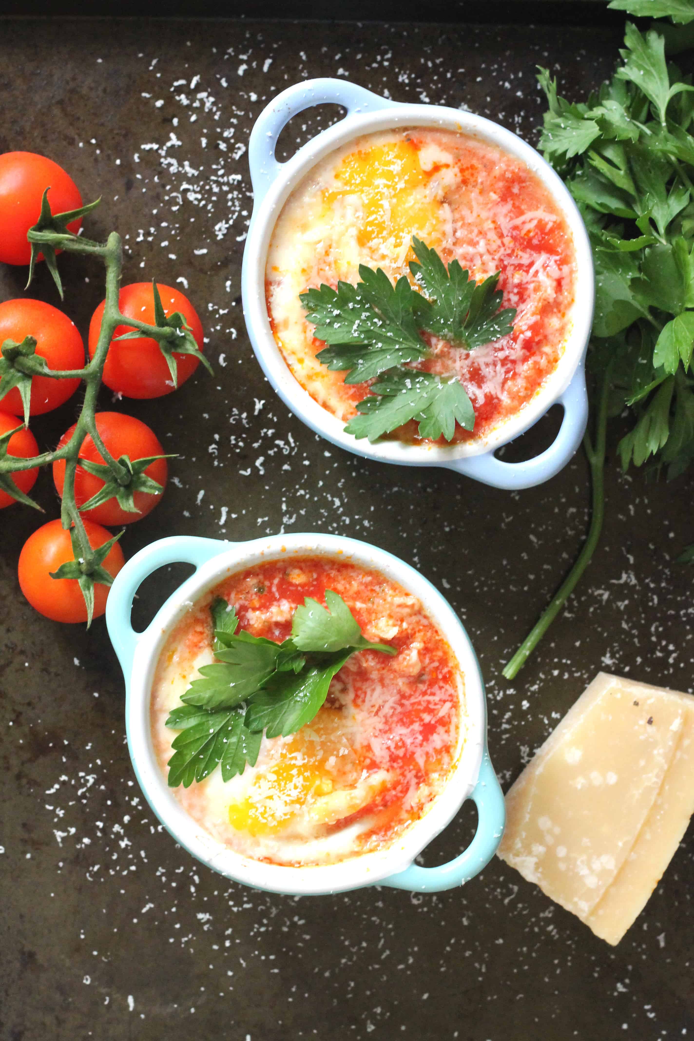 baked eggs with tomato and chorizo in individual white ramekins on a black background next to cherry tomatoes on the vine