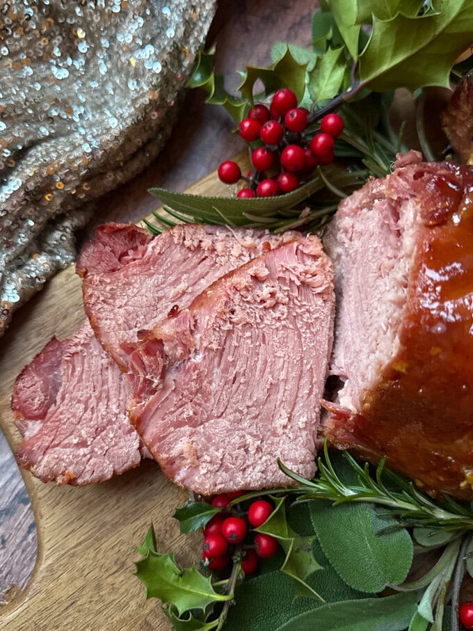 Sliced Slow Cooker Christmas ham sitting on a wooden chopping board on a bed of festive decor.