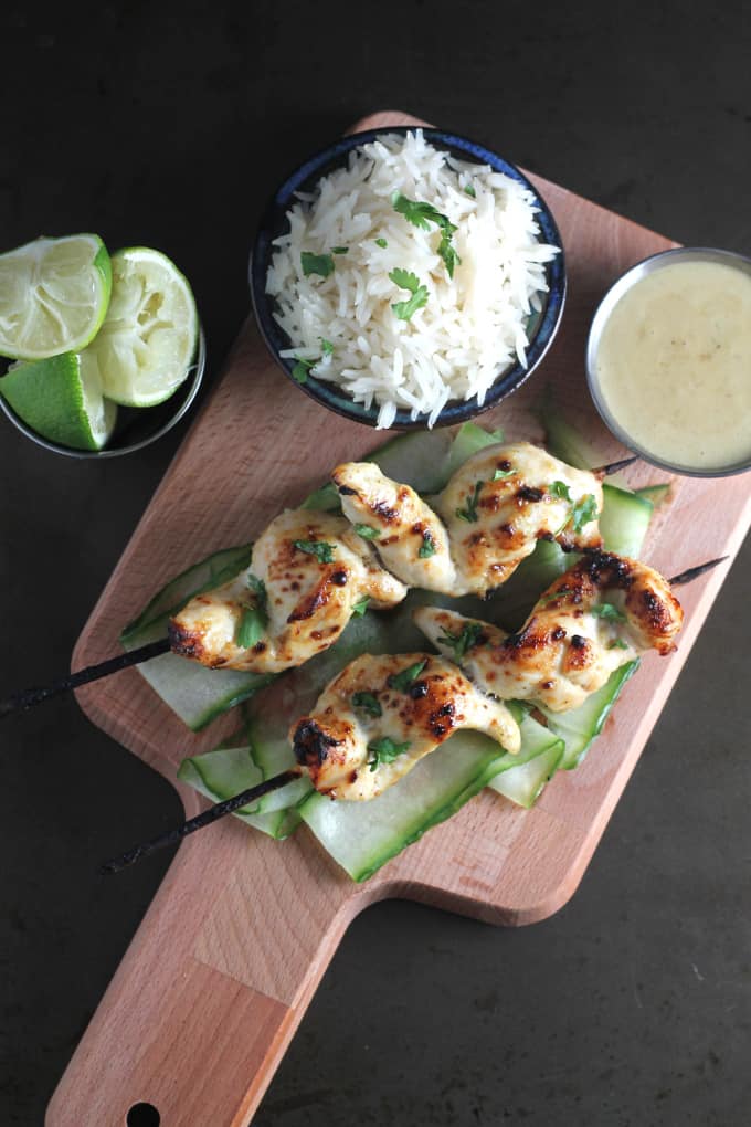 easy chicken satay skewers for kids served on a wooden chopping board with a small bowl of rice, some limes and a bowl of dipping sauce