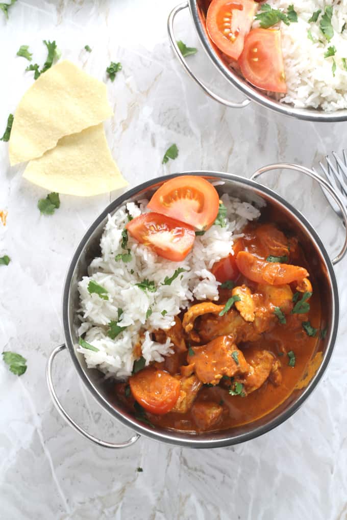 Chicken, Coconut & Tomato curry in a silver bowl with rice and two quarters of fresh tomato. On a marble background with a broken poppadom next to the bowl.