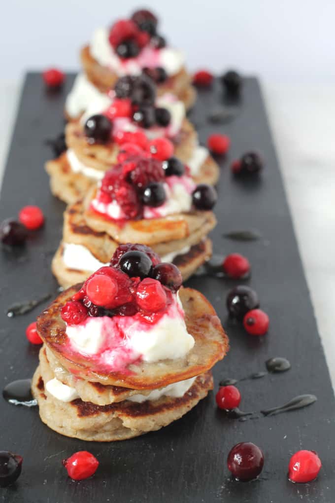 mini coconut banana buckwheat pancakes on a black slate serving tray topped with red berries and greek yoghurt