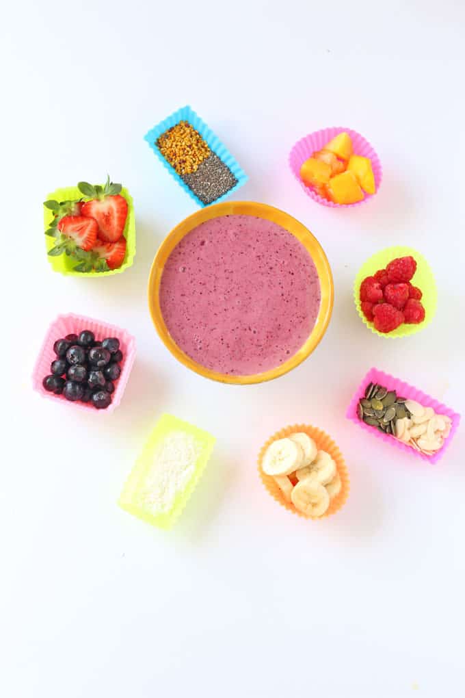 Fruit yogurt in a yellow bowl with a selection a multi coloured silicone muffin cases arranged around it containing nuts, seeds and berries