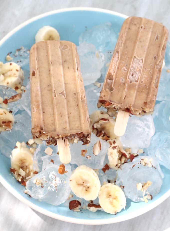 two peanut butter and banana popsicles in a blue bowl on top of ice cubes, banana slices and chopped nuts