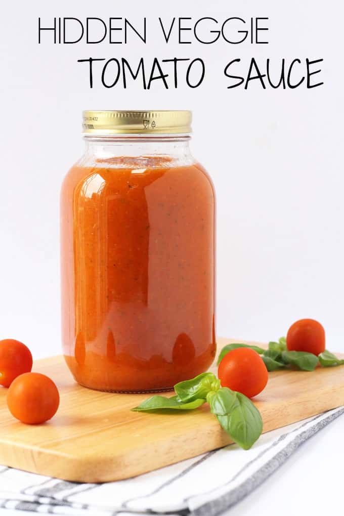 a jar of the hidden veggie tomato sauce on top of a chopping board scattered with cherry tomatoes and basil leaves