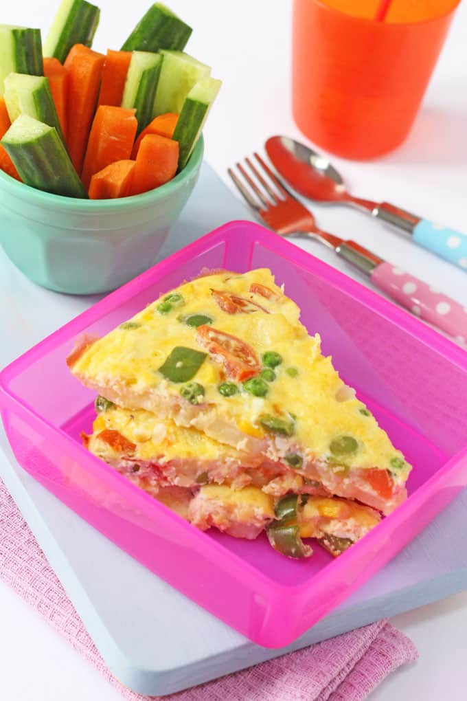 3 slices of lunchbox spanish omelette in a pink lunchbox on blue board. Served next to a blue ramekin with carrot and cucumber batons.