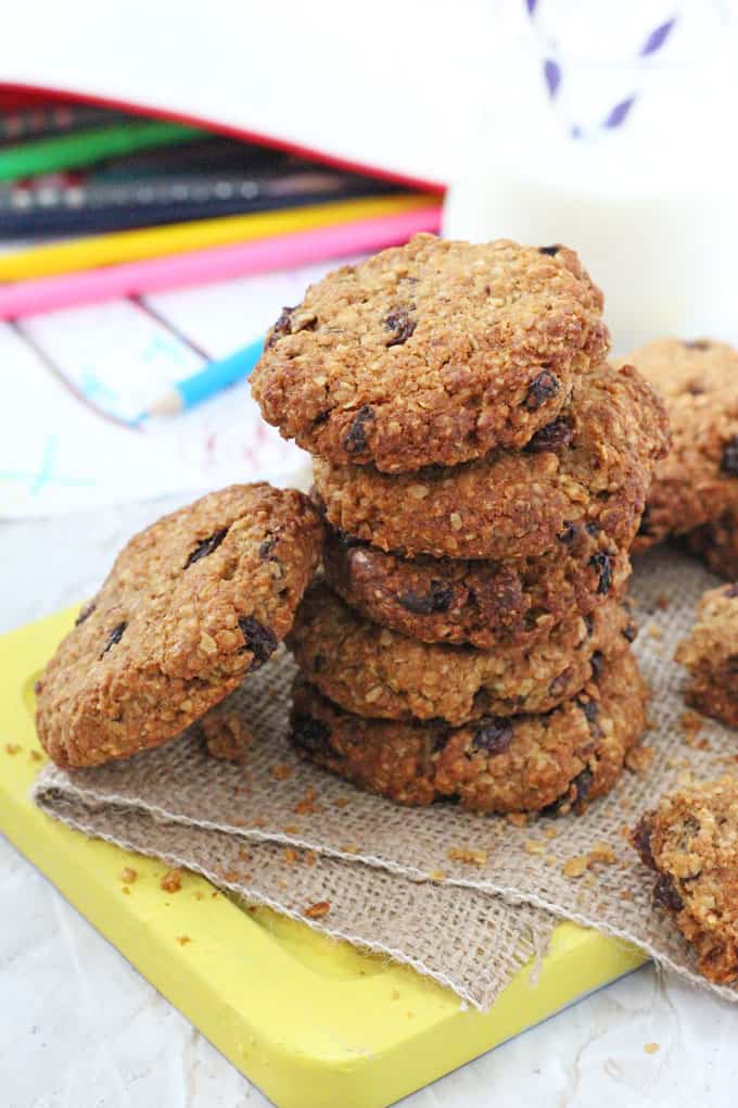 a pile of Oat & Raisin Cookies on top of a hessian mat on a yellow wooden chopping board with a pencil case in the background