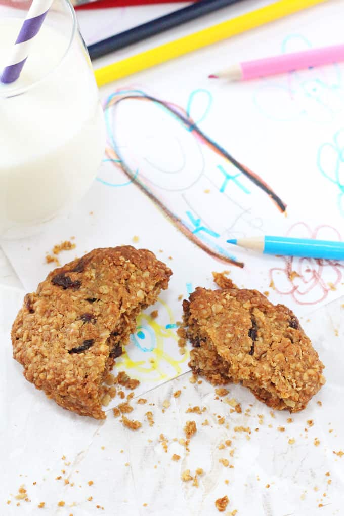An Oat & Raisin Cookie broken in half on top of a children's drawing next to a blue pencil and a glass of milk in the background
