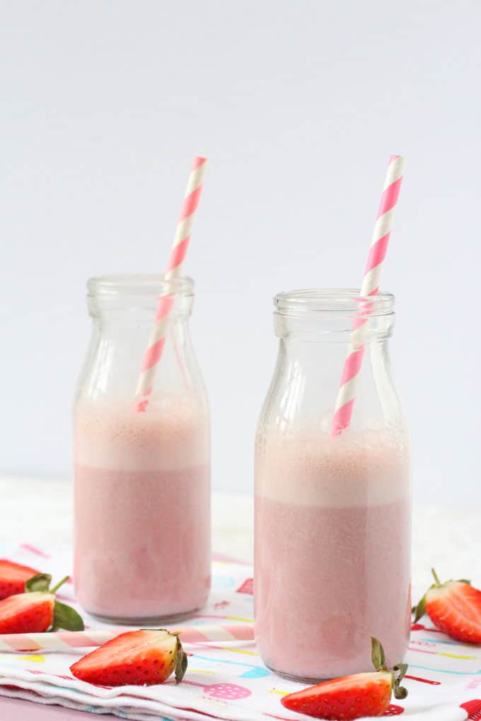 2 small milk bottles 3/4 filled with Strawberry Milk with a pink and white striped straw in each bottle. Surrounded with chopped strawberries