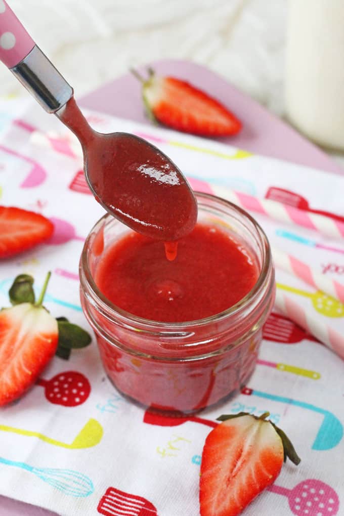 a teaspoon being dipped into a small jar of strawberry milk syrup. Sliced strawberries in the background an all sitting on top of white tea towel patterned with multi coloured utensils on it.