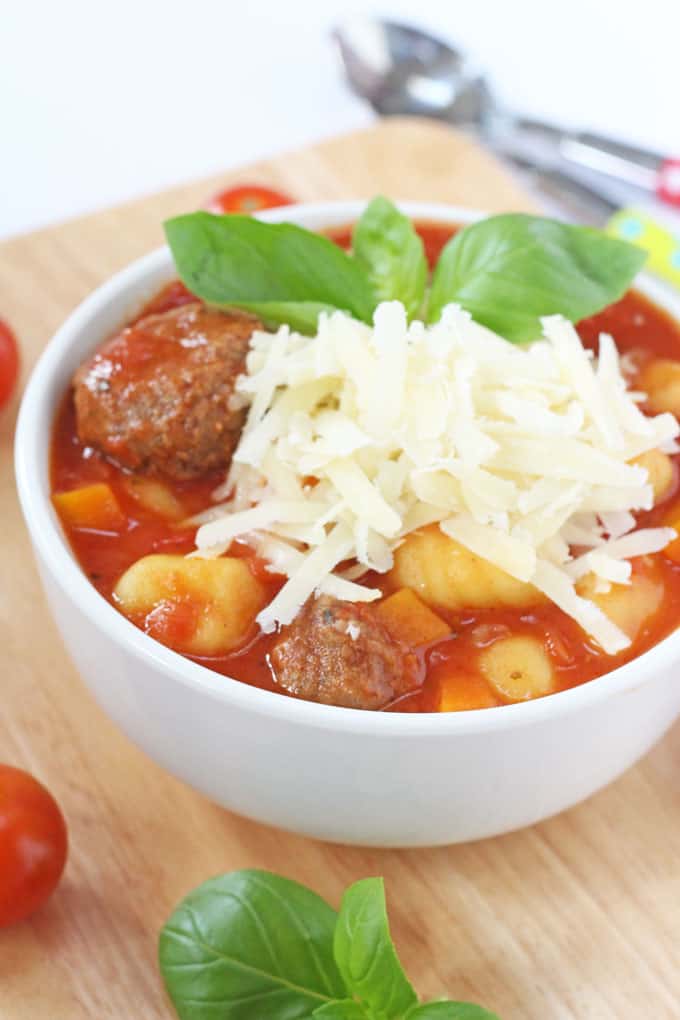 Slow cooker Italian meatball & gnocchi soup served in a white soup bowl, and garnished with grated cheese and basil leaves.
