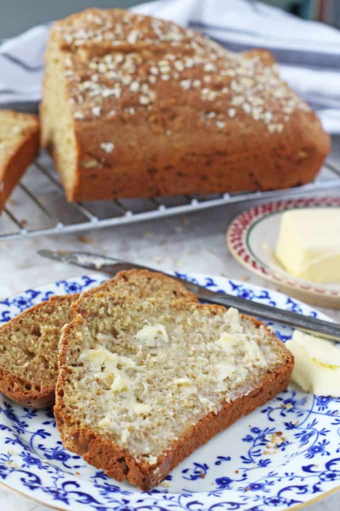 a loaf of easy irish soda bread on a cooling rack in the background Two slices of the buttered bread in the foreground, on a white and blue plate with a knife and butter resting on the side of the plate.