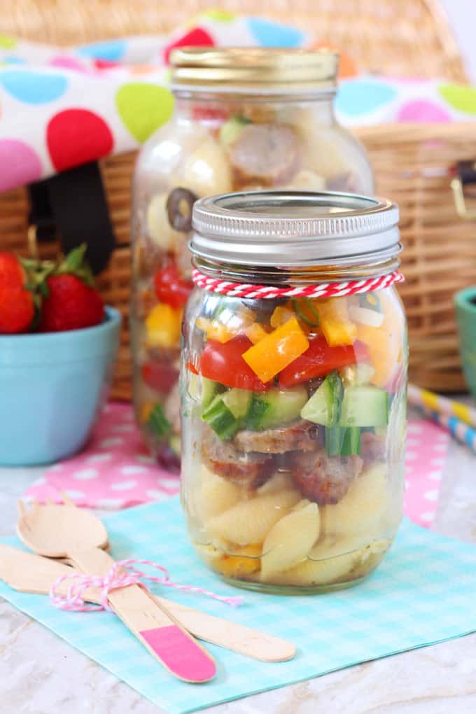 Italian sausage pasta salad in a mason jar in front of a picnic basket with wooden knives and forks to the side
