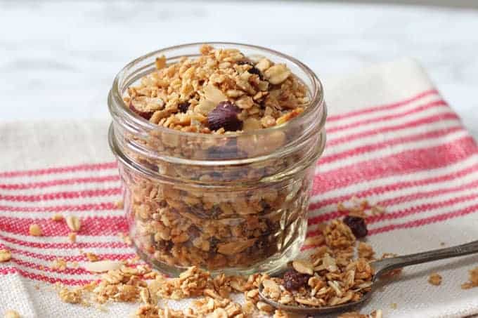 Easy Microwave Granola в стеклянной формочке на красно-белом полосатом кухонном полотенце с чайной ложкой.