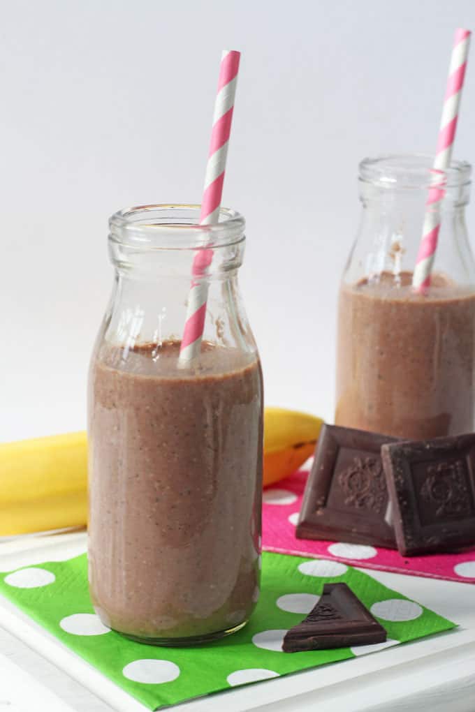 2 small milk bottles filled with the healthy chocolate chia shake sitting on pink and white and green and white polka dot napkins. There are 2 and a half squares of chocolate placed next to them and banana in the background.