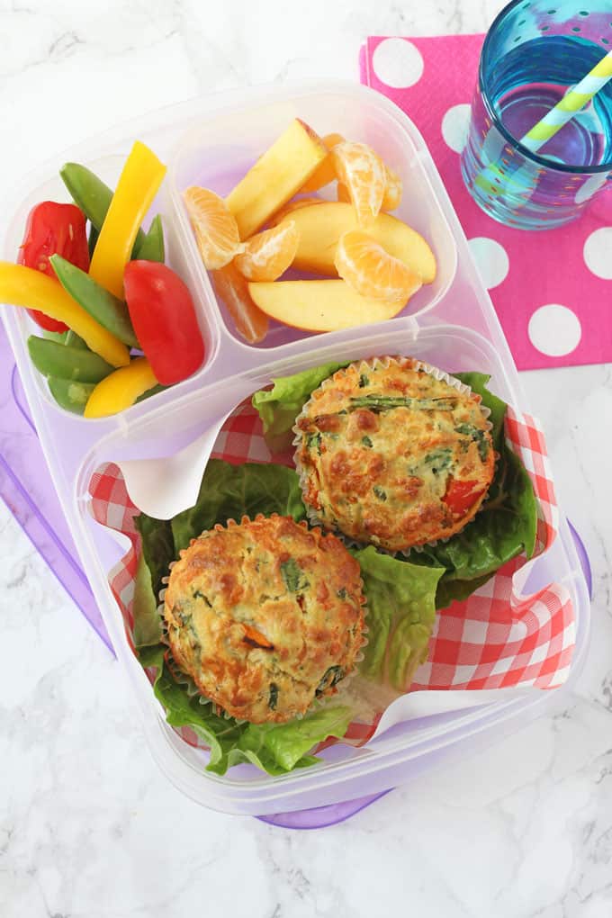 Savoury muffins in a compartment lunchbox, served with chopped peppers and cherry tomatoes and fresh orange and apple slices.