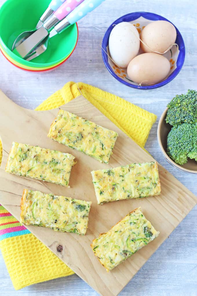 Broccoli and cheese frittata fingers on a wooden cutting board with the raw ingredients in the background.