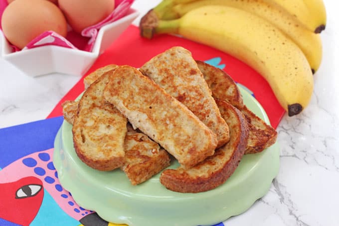 Baby French Toast or Eggy Bread cut into slices on a green serving plate with a bunch of bananas and eggs in the background.