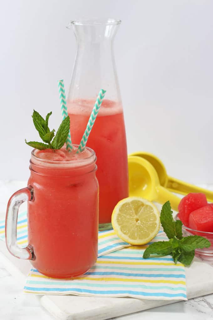 Watermelon Lemonade Served in a glass jar with a handle topped with a sprig of mint and a turquoise straw. In the background there is a jug of the watermelon lemonade, a bowl of watermelon chunks, a yellow lemon squeezer, half a lemon and a sprig of mint.