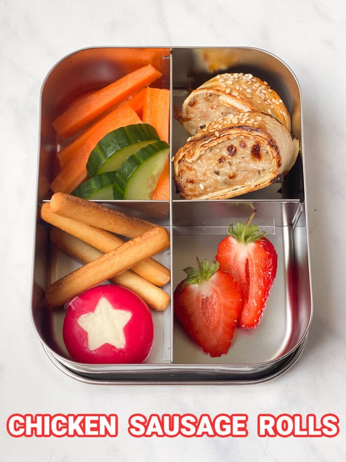 2 chicken sausage rolls in a stainless steel compartment lunchbox with mini breadsticks and fresh chopped fruit and veggies.