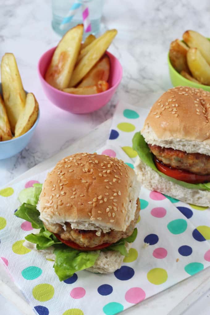 Chicken and apple burgers, served in sesame seed topped burger buns and garnished with a slice of tomato an lettuce leaves.