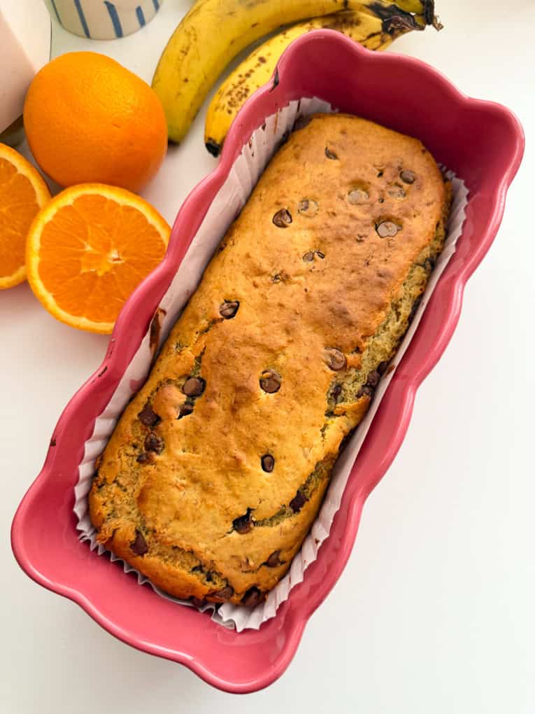 Easy Chocolate Orange Banana Bread (Healthy & Kid-Friendly) in a pink ceramic loaf tin with bananas and freshly cut oranges in the background.