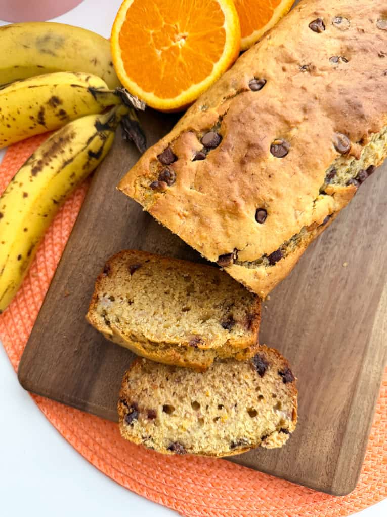 Easy Chocolate Orange Banana Bread (Healthy & Kid-Friendly) sliced on a wooden chopping board with bananas and orange halves in the background.