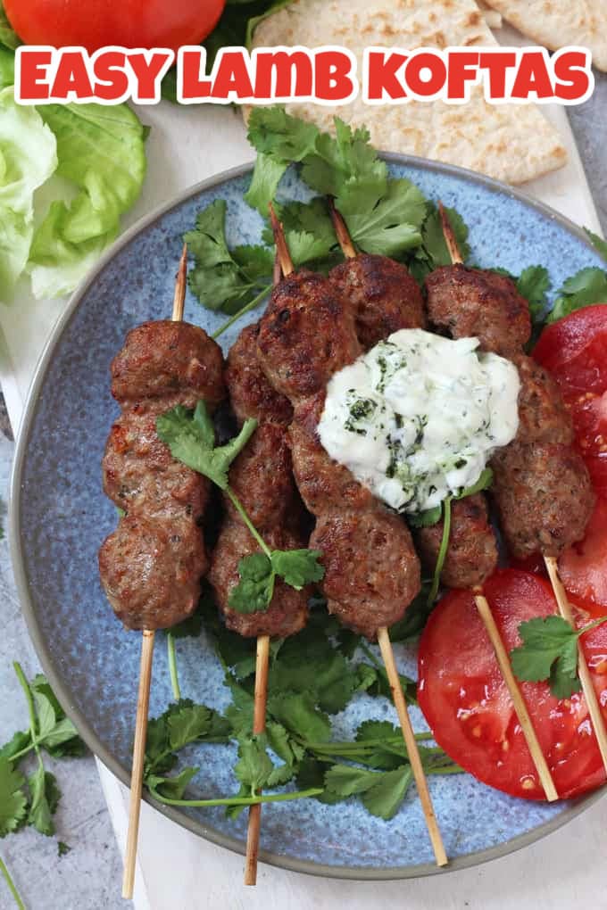 5 Lamb koftas on wooden skewers on a blue plate garnished with tomato slices, coriander and a dollop of tzatziki.