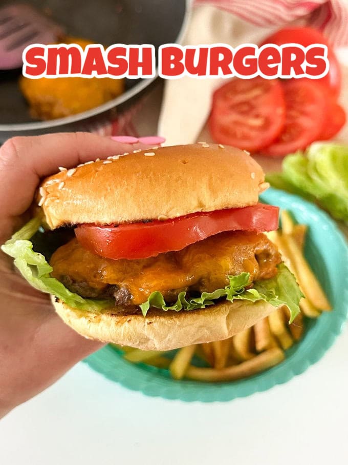 A hand holding a burger in a seeded bun with melted cheese, lettuce leaves and a slice of tomato inside.