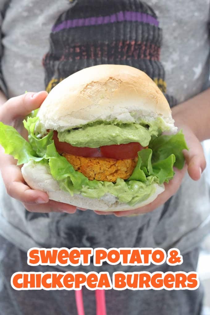 A child holding a burger bun containing a sweet potato and chickpea burger, lettuce leaves, a slice of tomato and guacamole.