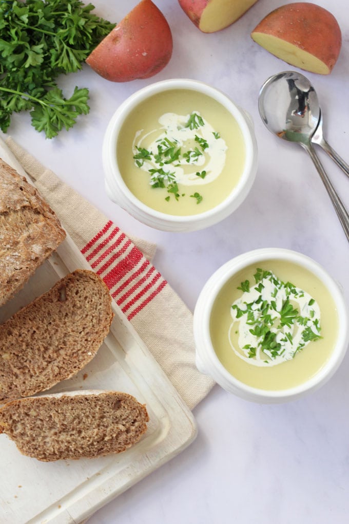 2 bowls of Cream Of Potato Soup in white bowls, garnished with creme fraiche and chopped herbs with a freshly cut loaf of bread on the side.