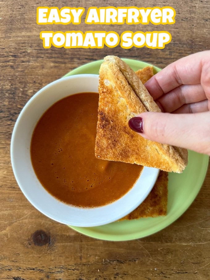 A single serving of air fryer tomato soup in a white bowl on top of a small greed side plate with a toasted sandwich being dipped into it.