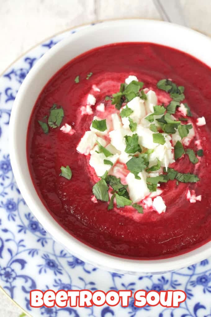 Beetroot & Carrot Soup served in a white bowl on top of a blue and white chintz plate and garnished with crumbled feta and freshly shopped herbs.