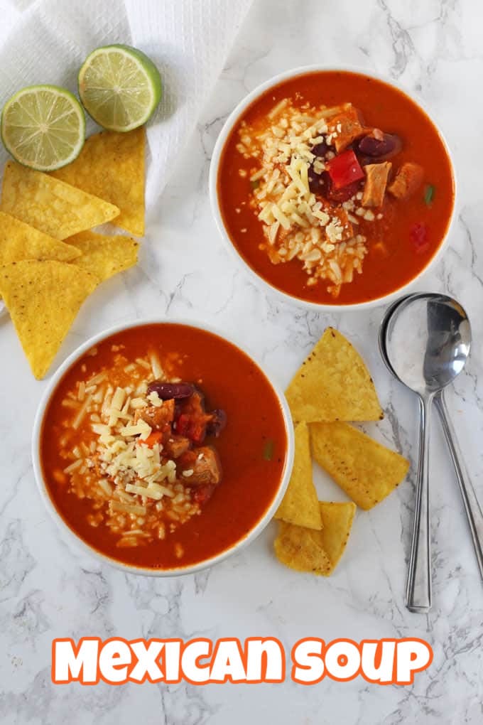 Two bowls of Mexican Soup on a marble surface with two soups spoons, some corn tortilla chips and two lime halves.