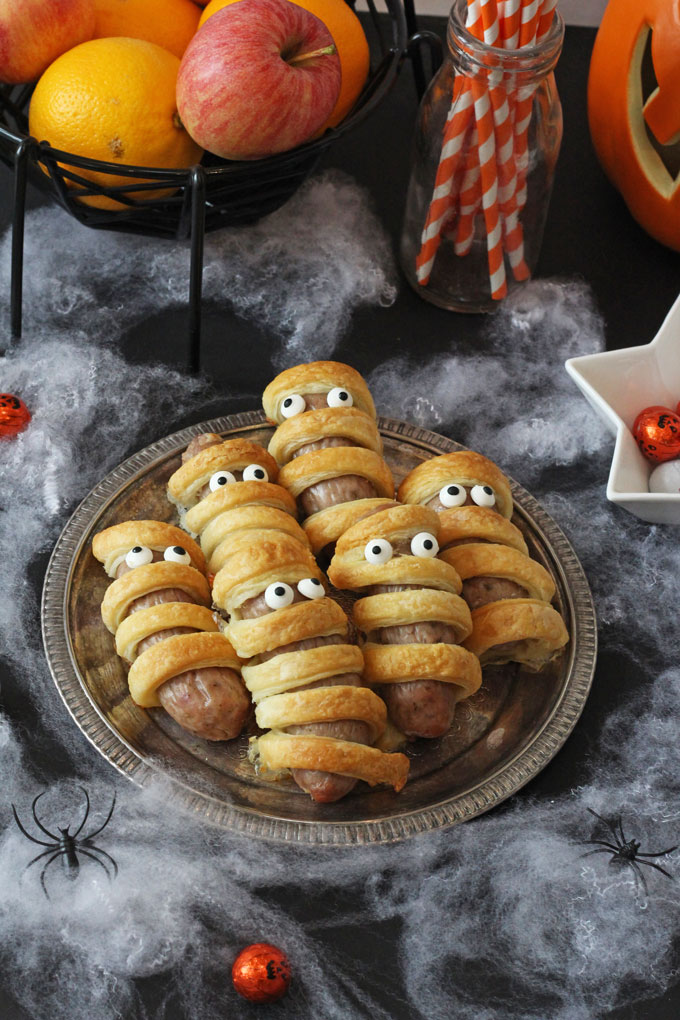 Fun Halloween Food Ideas For Kids -6 Sausage Mummies on a silver platter  with Halloween decorations in the background.