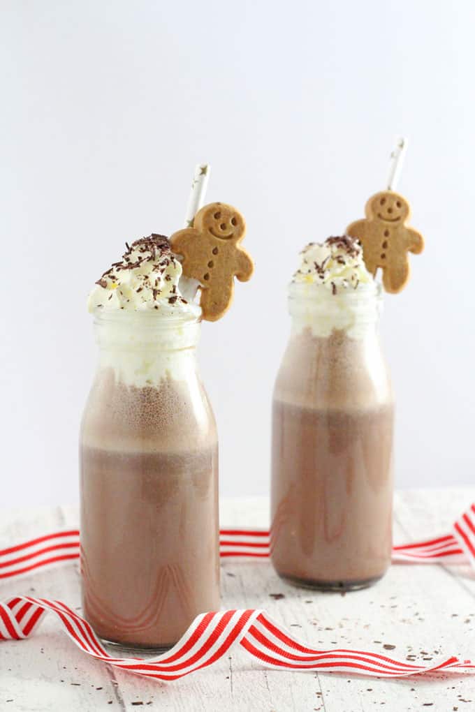 Frozen gingerbread hot chocolate in two small glass milk bottles, topped with whipped cream and chocolate shavings with a straw and a small gingerbread man in each.