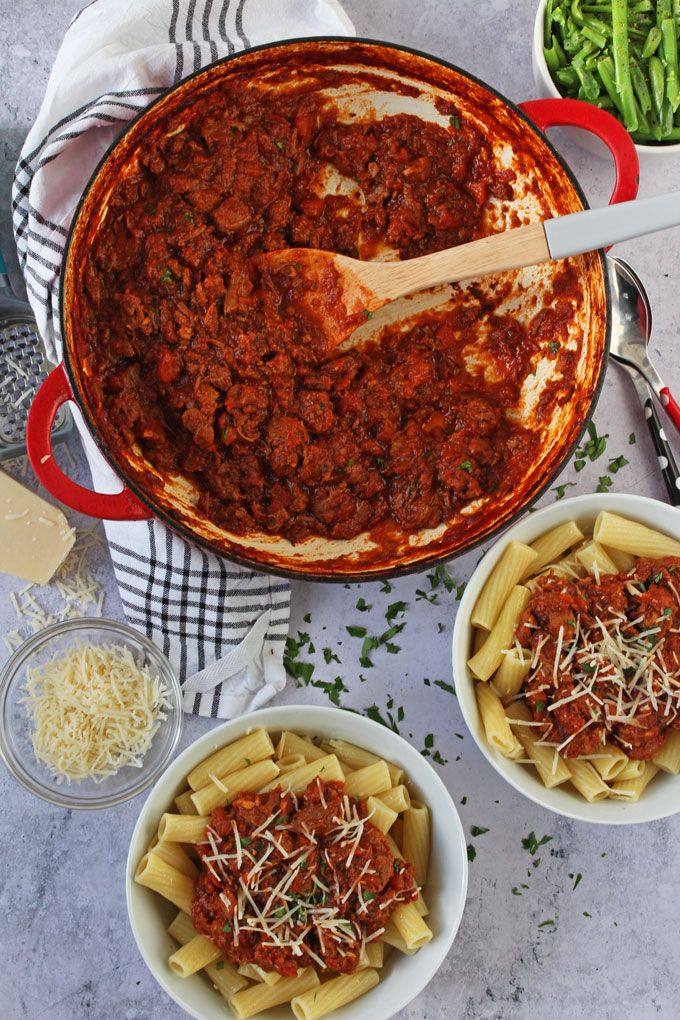 Slow Cooked Lamb Ragu sauce in a large red pot, with two bowls of the ragu served with cooked pasta in white bowls topped with grated parmesan