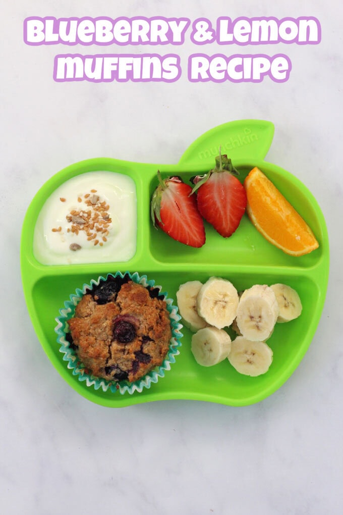 A Blueberry & Lemon Muffin on a green Munkin divided plate with sliced banana, halved strawberries, a wedge of orange and a small portion of Greek yogurt and seeds.