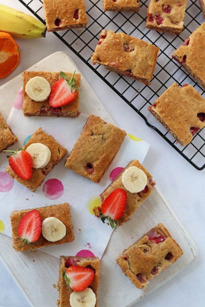 Strawberry & Banana Cake Bars topped with sliced banana and strawberry slices on a white chopping board with polka dot napkins.