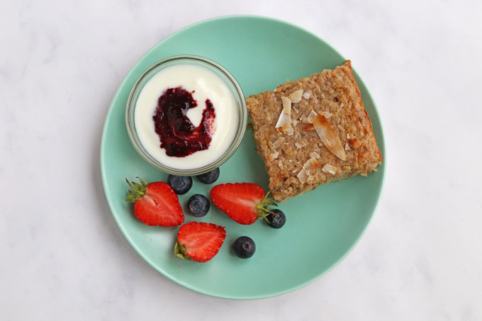 Coconut and almond breakfast bar, cut into a square and served on a turquoise plate with a glass ramekin with Greek yogurt and jam. With fresh strawberries and blueberries as a side dish.