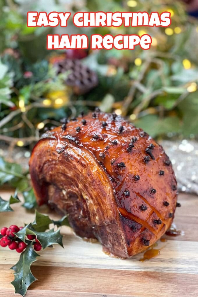 A Christmas Ham with a sticky glaze and studded with cloves sat on a wooden chopping board with fresh holly and twinkly lights in the background.