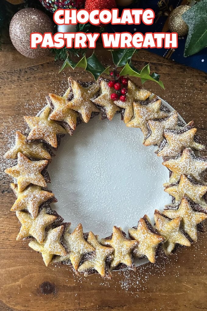 Chocolate Puff Pastry Wreath on a white plate dusted with icing sugar with fresh holly and Christmas baubles in the background.