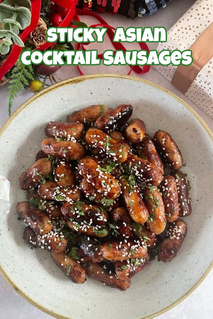 Stick Asian Cocktail Sausages garnished with sesame seeds and chopped herbs in a brown speckled bowl with Christmas decorations in the background.