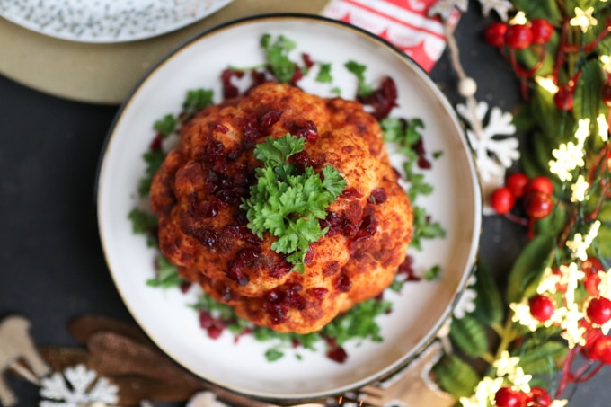 a whole roasted cauliflower on a white serving plate, garnished with fresh parsley and dried cranberries