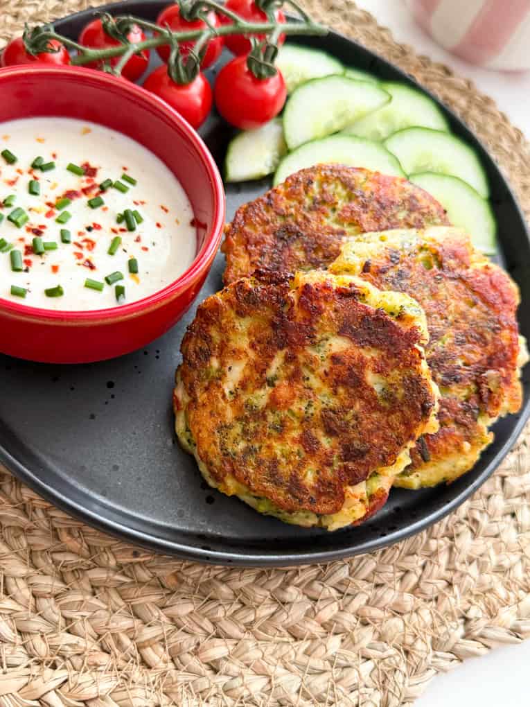 Chicken & broccoli fritters served on a black round plate with sliced cumber and a small red bowl filled with a dipping sauce.