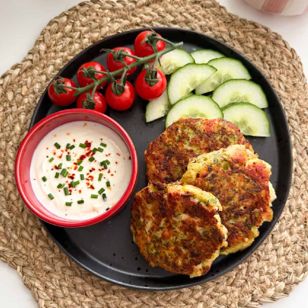 Chicken and Broccoli Fritters served on a black plate with cherry tomatoes, sliced cucumber and a ramekin of sauce.