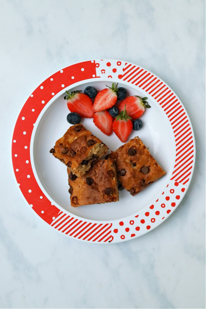 3 squares of chocolate chip cake bars on a red and white plate with some fresh fruit